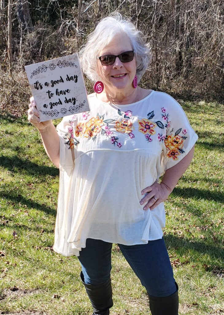 Judi in a embroidered flower shirt holding a sign reading Its a good day to have a good day